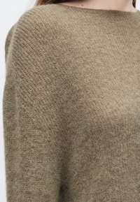 Pulover verde măsliniu, tricotat texturat, cu croială lejeră și decolteu larg, evidențiind un material moale și pufos, cu modele orizontale subtile.