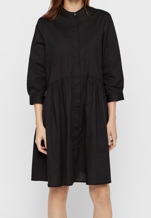 Kobieta ubrana w luźną, czarną sukienkę z długim rękawem, wysokim kołnierzem i marszczonym paskiem w talii, stojąca na jednolitym tle.