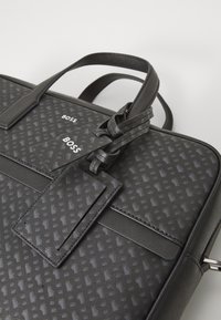 Sacoche en cuir noir pour ordinateur portable avec un motif géométrique texturé, deux poignées supérieures et une étiquette de bagages amovible arborant le logo de la marque.