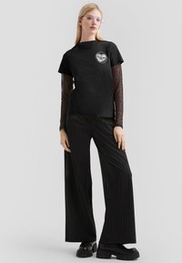 Schwarzes T-Shirt mit kurzen Ärmeln und grafischem Logo, übereinander getragen mit einem gemusterten Langarmoberteil. Kombiniert mit schwarzen Hemdstreifen-Hose mit weitem Bein und klobigen Stiefeln.