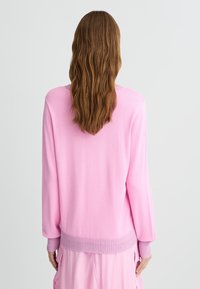 Maglione rosa a maniche lunghe con polsini e orlo a costine. Tessuto morbido, vestibilità ampia e design a scollo a V. Indossato sopra un indumento rosa.