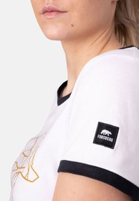 Camiseta blanca con ribetes negros y un diseño geométrico en dorado. Presenta una etiqueta negra con la palabra "FORSBERG" y un logo de oso en la manga.