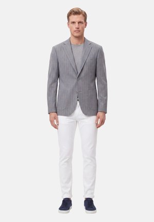 Uomo in piedi che indossa un blazer grigio a righe sottili, camicia grigio chiaro, pantaloni bianchi e sneakers blu scuro su uno sfondo bianco uniforme.