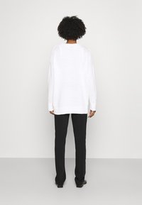 Maglione bianco a maglia con una vestibilità comoda, polsini e orlo a coste, abbinato a pantaloni neri e calzature minimal, visto da dietro.