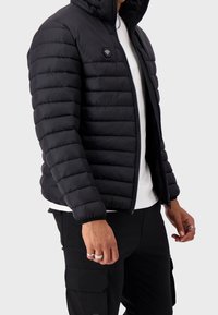 Giacca a vento nera con chiusura a zip, dotata di isolamento trapuntato orizzontale, colletto alto e patch con logo sul petto.