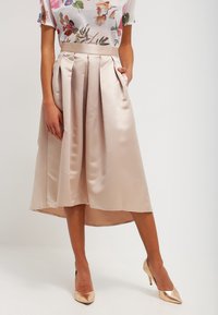 Satinová midi sukně v jemné béžové barvě s plisováním, se bočními kapsami a asymetrickým lemem, doplněná květinovým potiskem a tělovými podpatky.