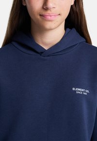 Persona joven con cabello largo castaño que lleva una sudadera con capucha de color azul marino con el texto "ELEMENT CO. SINCE 1992" en el pecho.