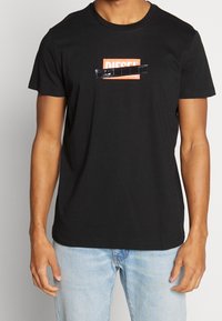 Camiseta de algodón negra con un gráfico rectangular naranja que tiene el texto blanco "DIESEL" y un acento negro en el medio. Mangas cortas, cuello redondo.