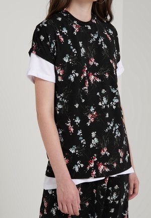 Woman wearing layered black floral top over white short sleeves and matching floral pants, standing against plain background.