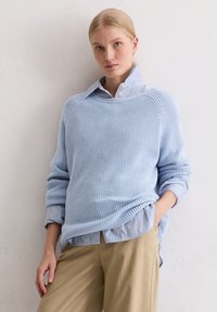 Hellblauer gestrickter Pullover mit Rippstruktur, über einem gestreiften Hemd mit Kragen getragen. Kombiniert mit beigen Hosen, die eine lässige Passform zeigen.