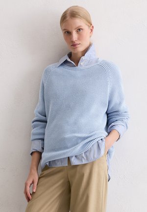 Hellblauer gestrickter Pullover mit Rippstruktur, über einem gestreiften Hemd mit Kragen getragen. Kombiniert mit beigen Hosen, die eine lässige Passform zeigen.
