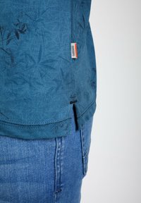 Chemise bleu à manches courtes avec un motif discret de feuilles, fentes sur les côtés et une étiquette de logo dans l'ourlet, associée à un jean en denim bleu clair.