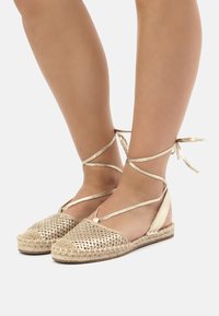 Espadrilles plates tissées en or avec une semelle en jute tressé et des liens à la cheville. Présente un motif en maille et une finition métallique lisse sur le dessus.