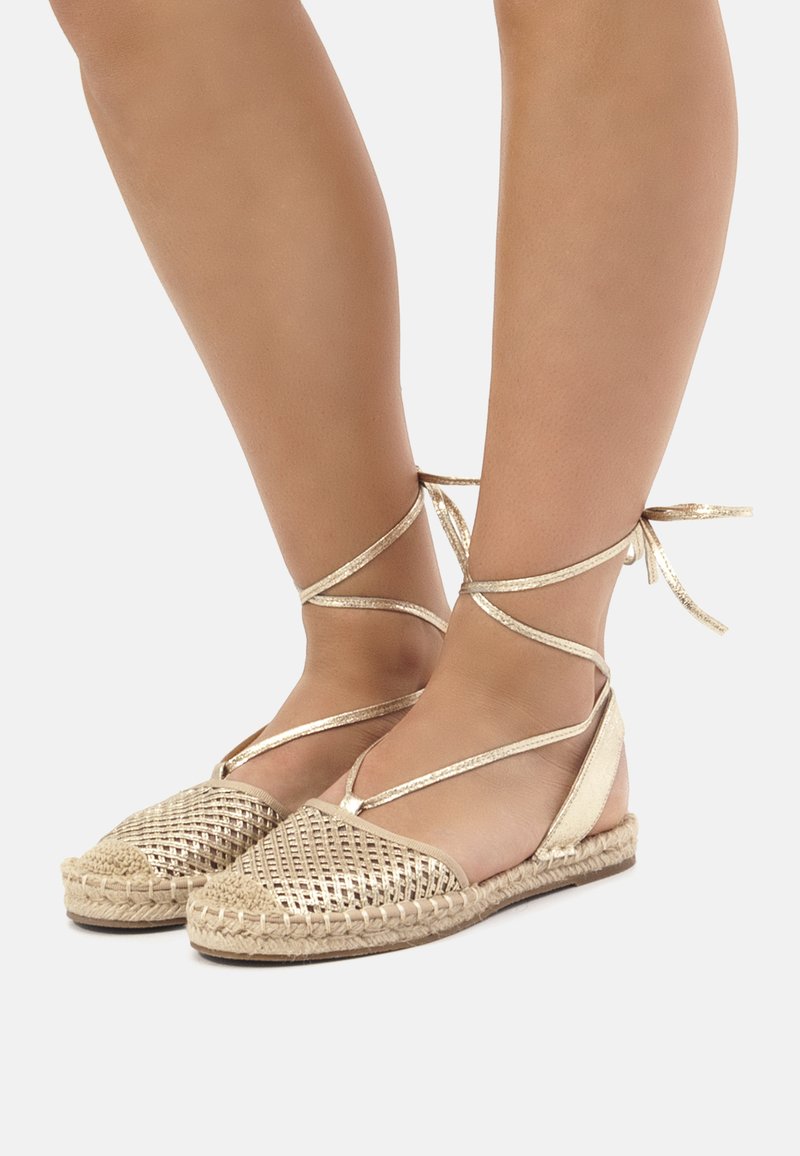 Espadrilles plates tissées en or avec une semelle en jute tressé et des liens à la cheville. Présente un motif en maille et une finition métallique lisse sur le dessus.