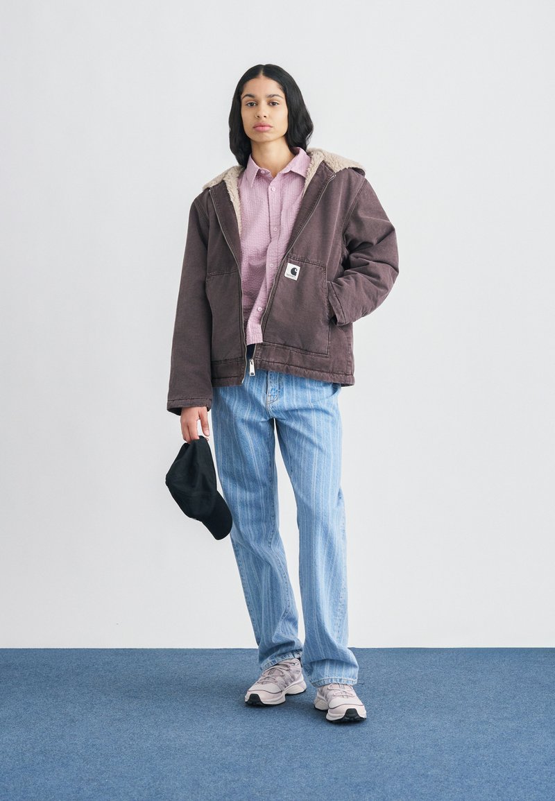 Brown insulated jacket with beige fur collar, light pink checked shirt, blue striped jeans, black cap in hand, and gray sneakers.