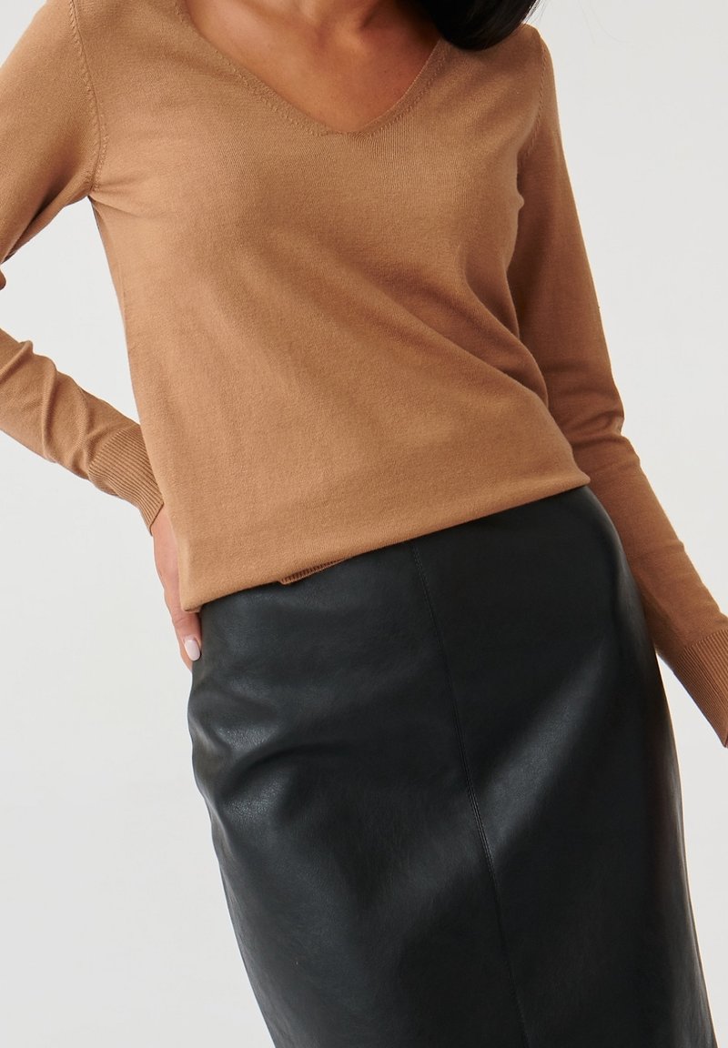 Femme portant un pull à manches longues couleur beige avec un décolleté en V et une jupe en cuir noire taille haute, main posée sur la hanche, sur fond blanc.