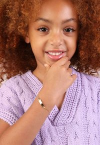 Gouden armband om de pols van een kind, met een glad, gepolijst oppervlak. De gebreide paarse trui heeft een kabelpatroon en een V-hals ontwerp.