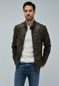 Giacca di pelle marrone con colletto alto, zip frontale e tasca sul petto. Indossata sopra un maglione bianco a trama, abbinata a jeans blu.