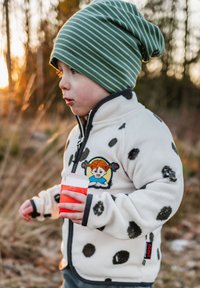 Valkoinen fleece-takki, jossa on mustia pallokuvioita, vetoketjukiinnitys, ja jossa on värikäs brodeerattu tägi sekä tummanvihreä raidallinen pipo. Lapsi pitää kädessään punaista juomaa.