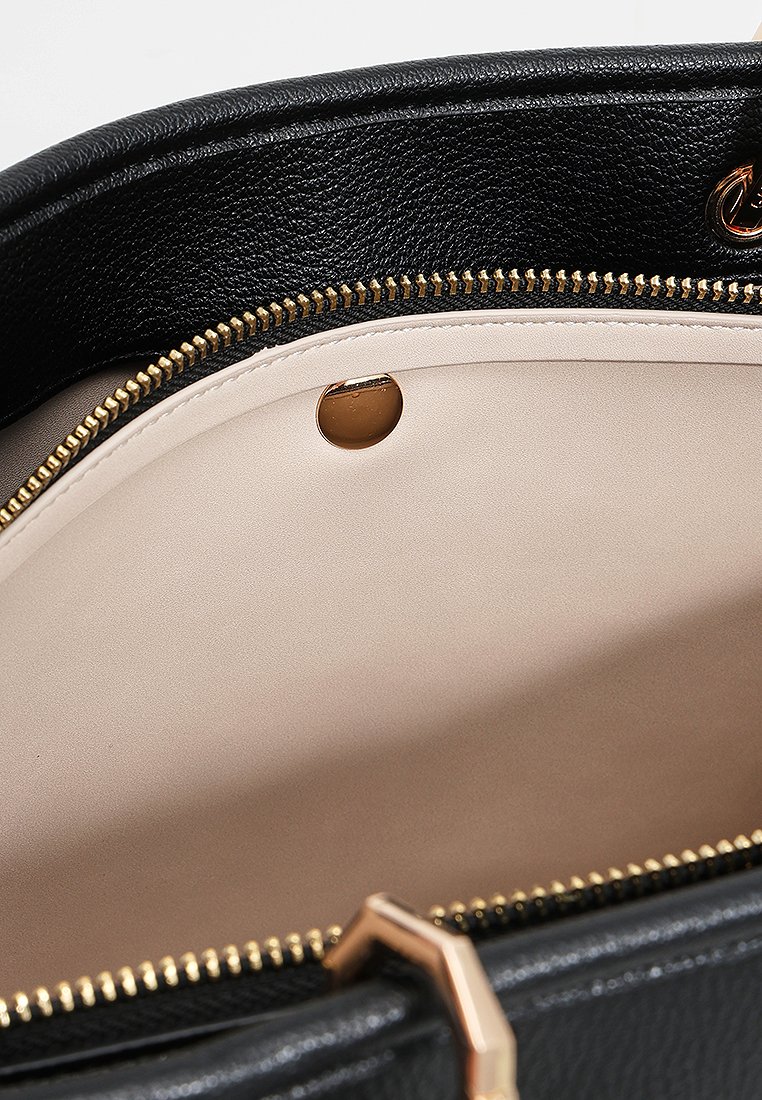 Interno di una borsa nera con texture, caratterizzato da una fodera beige, una cerniera dorata e un dettaglio con bottone a pressione circolare dorato.