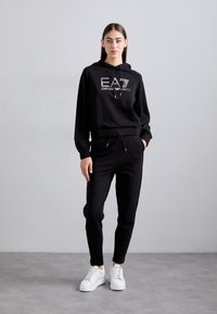 Jauna moteris stovi su juoda EA7 Emporio Armani džemperiu, suderintomis juodomis kelnėmis ir baltais sportbačiais prieš pilką fono sieną.