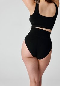 Frau trägt schwarze hochgeschnittene Unterwäsche und schwarzen Sport-BH, steht mit teilweise gedrehtem Rücken und erhobenen Armen.