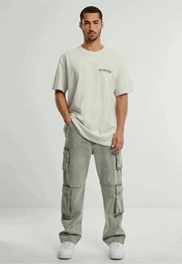 Mies seisoo yllä vaaleahkon ylimitoitetun t-paidan ja vaaleanharmaiden cargo-housujen kanssa, jalassa valkoiset tennarit yksinkertaista taustaa vasten.