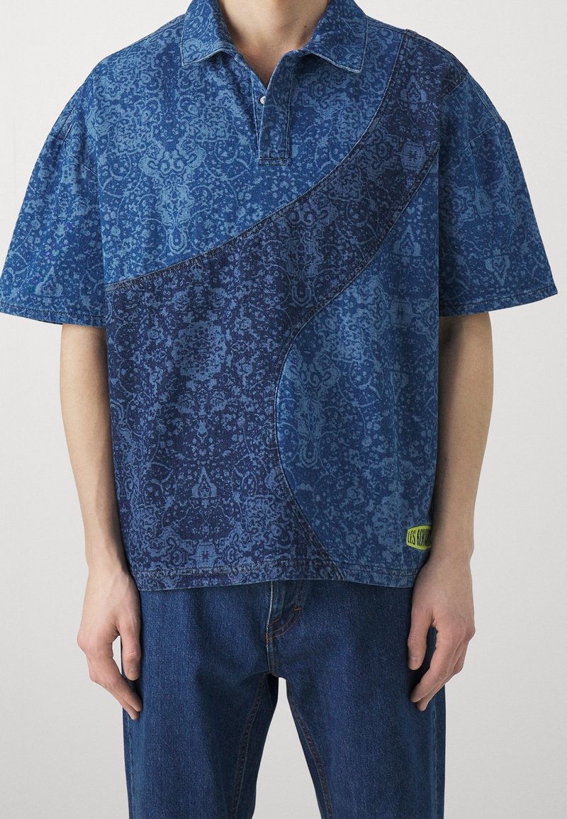 Homme portant un polo bleu à manches courtes avec un panneau incurvé plus foncé et un jean bleu sur un fond uni.