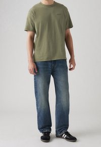 Man i olivgrön kortärmad t-shirt från Levi's, urblekta blå jeans och svarta Adidas-sneakers med vita ränder, stående på grått golv.