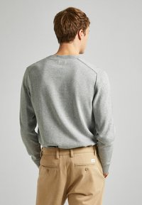 Grauer gestrickter Pullover mit langen Ärmeln und einem gerippten Halsausschnitt, kombiniert mit hellbraunen Hosen, zeigt die Rückansicht und minimale Details.