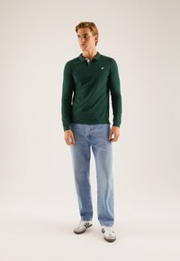 Langermet grønn polo skjorte med hvit krage, parret med lyseblå jeans og svart-hvite joggesko. Enkelt design, ingen mønstre.