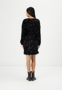Vestido preto com lantejoulas, mangas longas, corte solto e decote arredondado, combinado com botas pretas até o tornozelo. Superfície texturizada e brilhante.