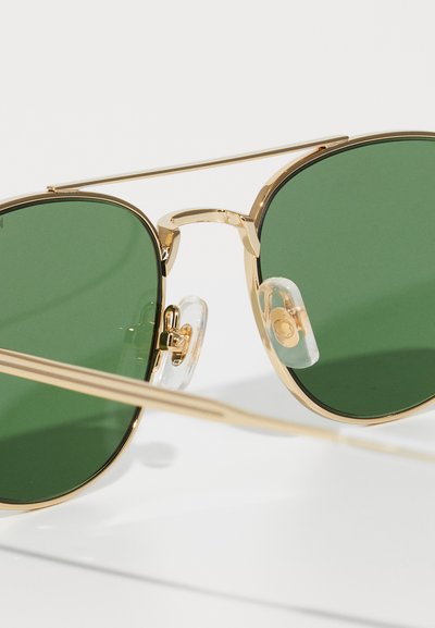 Ochelari de soare aviator aurii cu lentile verzi, cadru din metal, tampoane nazale clare și un design elegant al punții. Suprafață reflectorizantă și temple alungite.