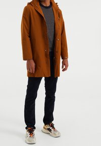 Ruosteenvärinen, vedenkestävä takki hupulla, painonapeilla ja sivutaskuilla, yhdistetty kuvioituun neuleeseen, tummiin housuihin ja urheilullisiin kenkiin.