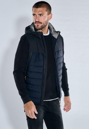 Navy puffer vest with a gray hood, side pockets, and a zipper closure, layered over a dark knit sweater, worn with black jeans.