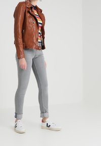 Person med en brun läderjacka, regnbågsrandig tröja, ljusgrå jeans med uppvikta ben och vita sneakers med svarta detaljer.