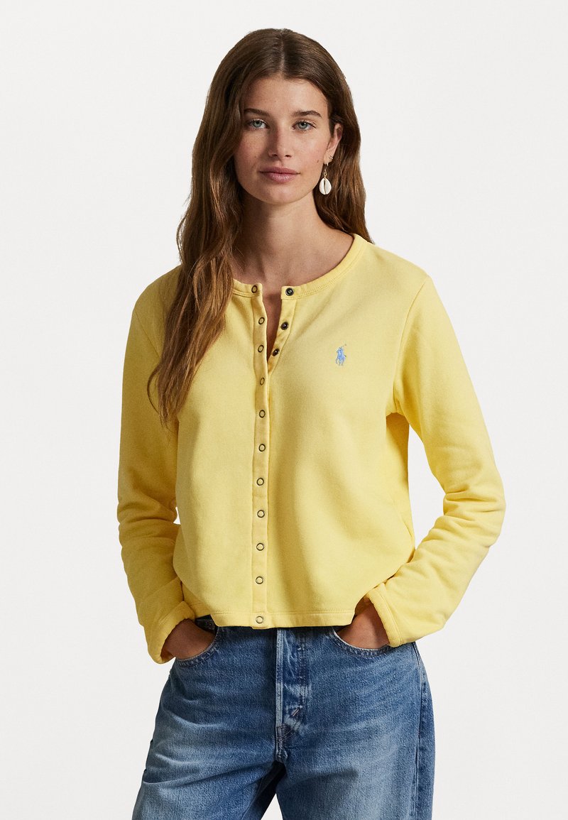 Yellow cotton sweater with a round neck, long sleeves, and snap buttons. Features a small blue logo on the chest. Worn with light blue jeans.