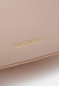 Rožinė tekstūruota oda su aukso spalvos iškilniu "Coccinelle" logotipu. Lygi kraštai ir švelnus paviršius pagerina taktilinę kokybę.