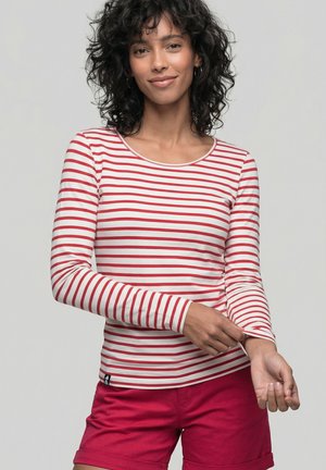 Frau mit lockigem dunklem Haar, die den Ärmel eines rot-weiß gestreiften Langarmshirts hochkrempelt, trägt rote Shorts, neutraler Hintergrund.