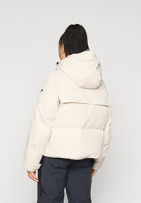 Πουπουλένιο μπουφάν σε ανοιχτό μπεζ χρώμα με κουκούλα, με οριζόντια επένδυση, φάσα στην πλάτη και λογότυπο στον αριστερό ώμο.