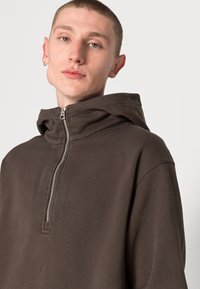 Braune Zip-Hoodie aus weichem Stoff, mit hohem Kragen und einem Frontreißverschluss. Einfaches Design ohne zusätzliche Muster oder Akzente.