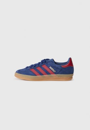 Sinine sametist sneaker punaste külgtriipude ja kuldse "Gazelle" brändimisega. Omab kummist kummipealse ja pehmendusega kaela mugavuse tagamiseks.