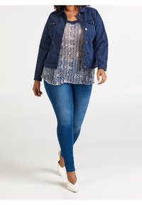 Mujer caminando con chaqueta azul, blusa estampada, jeans ajustados azules y tacones blancos en un fondo blanco.