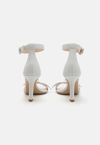 Tacones abiertos blancos con un delgado tacón de aguja, una correa alrededor del tobillo y una textura de cuero suave. Plantilla de color beige claro visible.