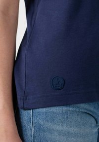 Primer plano de una persona que lleva una camiseta azul marino con un pequeño logo de JOTT cerca do la costura, combinada con unos jeans de denim azul claro.
