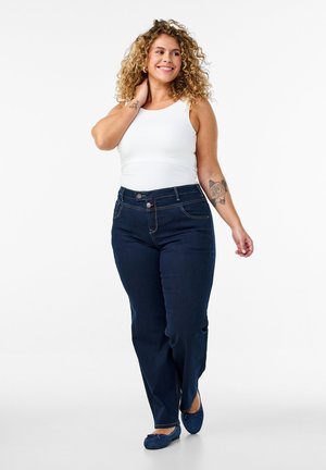Curly-haired woman in white sleeveless top and dark blue jeans with rose tattoo on arm, smiling and walking against white background.