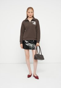 Giovane donna che indossa un pullover marrone con zip a quarto, una mini gonna nera lucida, delle ballerine rosse e tiene in mano una borsa marrone su uno sfondo bianco semplice.