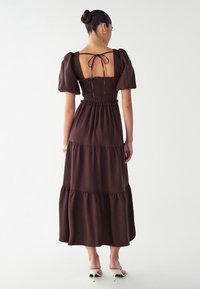 Robe maxi marron à volants avec des manches bouffantes et un corsage smocké. Présente un dos ouvert noué avec un ruban. Tissu lisse, design décontracté.