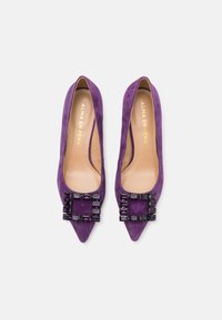 Tacones altos de ante púrpura con punta afilada, que presentan una gran hebilla de joya negra en la parte delantera. El interior tiene un forro beige.