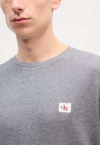 Γκρίζο textured t-shirt με στρογγυλή λαιμόκοψη και κοντά μανίκια. Διαθέτει μια μικρή λευκή ετικέτα με κόκκινο λογότυπο "K" στη αριστερή πλευρά.